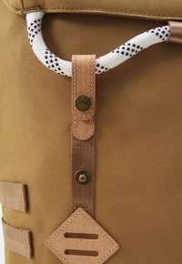 Sac à dos en tissu marron avec une poignée en corde blanche, des accents en cuir et un bouton-pression en laiton. Il comporte des boucles de sangle tissées et un patch en forme de losange.