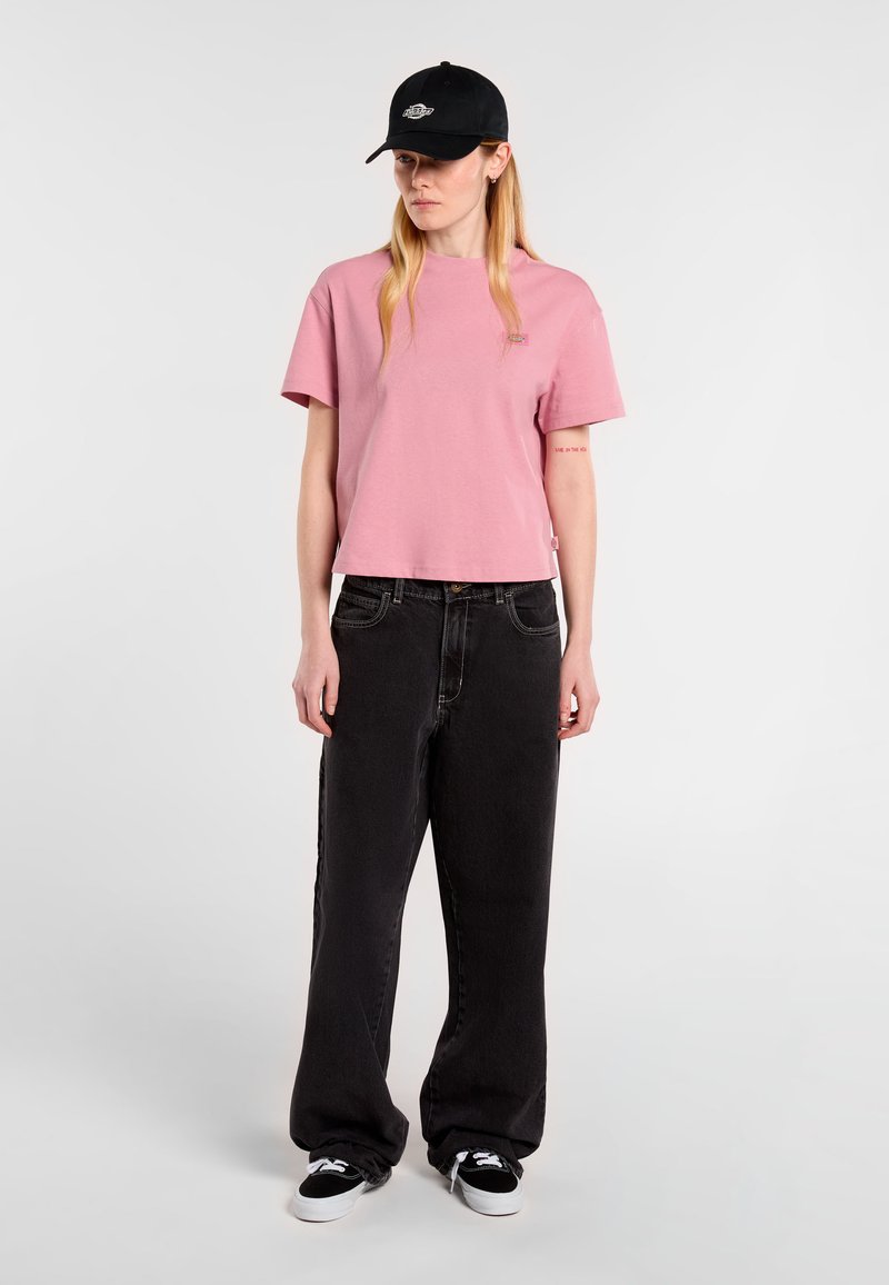 T-shirt corta a maniche corte color rosa, jeans neri a vita alta e a gamba larga, e cappellino nero con logo. Sneakers nere. Design semplice, look casual.