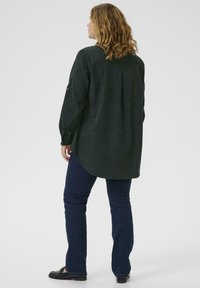 Tmavě zelená dlouhá rukávová košile z měkké tkaniny, s skladem na zádech, zaobleným lemem a detaily na knoflících na manžetách. Nosit s tmavými džíny.
