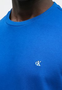 Bawełniany t-shirt w królewskim niebieskim kolorze z okrągłym dekoltem, z małym białym haftowanym logo „cK” na lewej stronie klatki piersiowej.