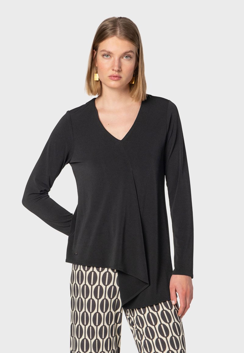 Svart långärmad topp med V-hals och asymmetrisk nederkant, tillverkad av en slät tyg. Kombinationen med mönstrade byxor med cirkulära mönster.