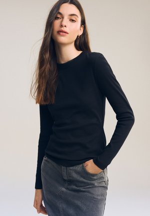 Donna con lunghi capelli castani, indossa una maglia nera a maniche lunghe e una gonna di jeans grigia, in piedi con una mano in tasca su uno sfondo neutro.