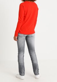 Rode longsleeve shirt met ronde hals, gecombineerd met strakke lichtgrijze jeans, met minimale details en witte sneakers.