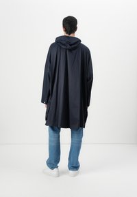 Mørkeblå hetteponcho laget av lett stoff med løse ermer, midt på låret lengde, kombinert med lyseblå jeans og hvite sneakers.