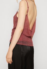 Donna che indossa un top senza maniche rosso scintillante con la schiena bassa scoperta e pantaloni di satin neri, mostrata di lato e di schiena.