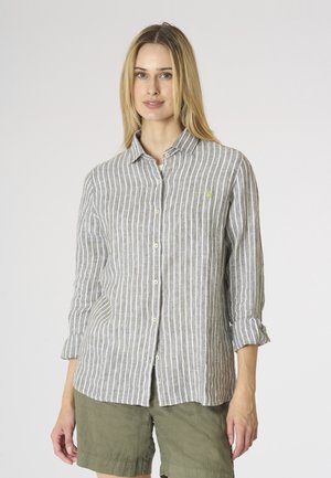Donna con camicia a righe verticali grigie e bianche abbottonata, maniche arrotolate e pantaloncini verde oliva, in piedi davanti a uno sfondo uniforme.