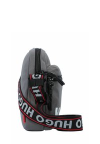 Bolsa de nailon gris con cierre de cremallera, que presenta una correa audaz en negro y rojo con el logo "HUGO" y detalles texturizados. Forma compacta y rectangular.