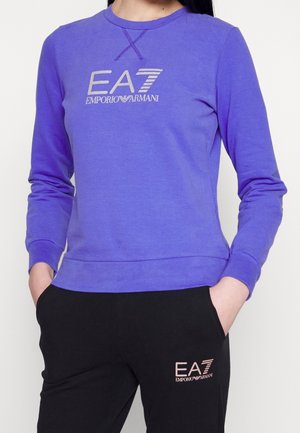 Personne portant une chemise à manches longues violette et un pantalon noir, tous deux avec des logos blancs Emporio Armani EA7, mains dans les poches.