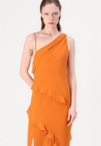 Orange, asymmetrisk klänning med en axelrem, har volanger längs fållen och en sidoklyvning. Slätt tyg med en flytande design.