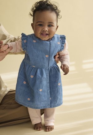 Bébé souriant en robe bleue avec broderie de fleurs et leggings roses, debout pieds nus tout en étant soutenu doucement.