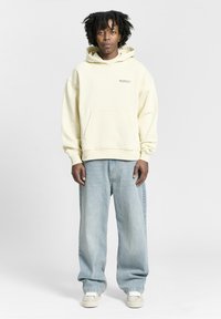 Hellgelbe, übergroße Kapuzenjacke mit einer Fronttasche und Logo auf der Brust, kombiniert mit hellblauen weit geschnittenen Jeans und weißen Sneakern.