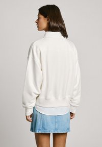 Sudadera blanca de gran tamaño con un cuello alto, puños y dobladillo de canalé, combinada con una falda de mezclilla azul clara. Textura suave.