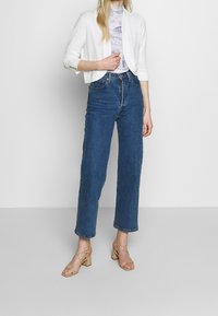 Cardigan bianco corto, top chiaro a fantasia, jeans blu a vita alta e sandali beige con tacco quadrato, con una vestibilità rilassata e gamba dritta.