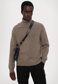 Hellbrauner Rollkragenpullover aus weichem Strickmaterial, kombiniert mit einer schwarzen Umhängetasche mit silbernen Akzenten und verstellbarem Schultergurt.