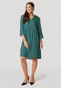 Grünes Kleid mit geometrischem Muster, V-Ausschnitt und Dreiviertelärmel. Das Material ist leicht, hat eine lockere Passform und einen fließenden Saum.