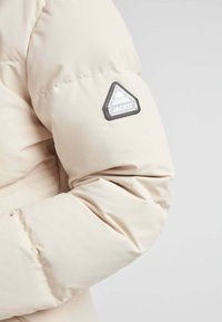 Manga de chaqueta hinchada beige con textura acolchada. Presenta un parche triangular negro con el texto blanco "JACKIT" y un diseño geométrico.