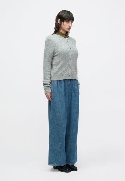 Ανοιχτό γκρι πλεκτή ζακέτα με κούμπωμα με κουμπιά, σε συνδυασμό με φαρδιά μπλε τζιν παντελόνια. Το ντύσιμο έχει άνετη γραμμή και απλό σχεδιασμό.