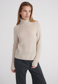 Beiger, weicher Strickpullover mit hochgezogenem Rippkragen und langen Ärmeln; ribbeliger Saum an der Taille. Kombiniert mit dunkelgrauen weiten Hosen.