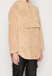 Pullover in pile beige, morbido e con cappuccio, con zip mezza lunghezza frontale e tasca a pattina. Presenta un design angolato e una tasca laterale con zip sul lato sinistro in basso.