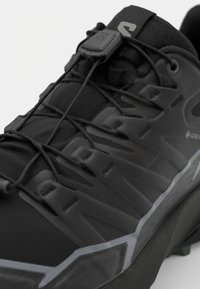 Zwarte sportieve schoen met een lichtgewicht bovenwerk, textuur op het oppervlak, geïntegreerd vetersysteem en rubberen zool met een kenmerkend profiel.