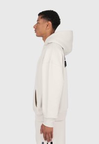 Young man wearing light beige oversized hoodie and matching sweatpants, standing in profile against a plain background.