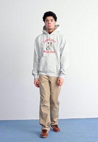 Sudadera gris con un gráfico de "Carhartt Wild Dogs", pantalones beige y zapatos marrones. La sudadera cuenta con un bolsillo frontal y puños acanalados.
