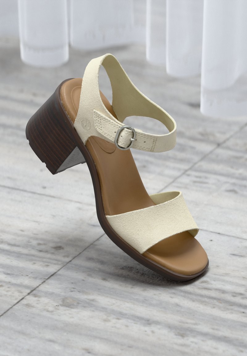 Μπεζ σανδάλι από σουέντ με ανοιχτά δάχτυλα, ρυθμιζόμενο λουράκι στον αστράγαλο και στιβαρό τακούνι από ξύλο, πάνω σε γκρι πλακόστρωτο δάπεδο.