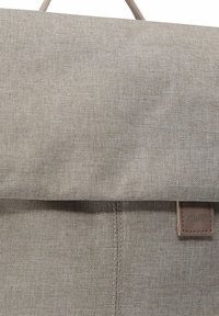 Mochila de tela gris con una solapa plegable, que presenta un parche de acento de cuero marrón. La textura es tejida y suave, con detalles de costura visibles.