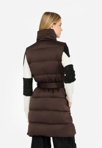 Gilet imbottito marrone isolato con colletto alto, vita cinturata e maniche testurizzate a contrasto bianco e nero. Superficie liscia e trapuntata.