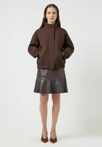 Chaqueta marrón acolchada con textura floral, cuello alto, bolsillos delanteros; combinada con una falda acampanada de cuero marrón oscuro y tacones puntiagudos.