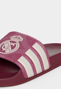 Sandale slide marron avec des rayures blanches et le blason du Real Madrid sur la sangle, semelle intérieure texturée et design à bout ouvert.
