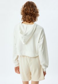 Gekürzter, weißer Hoodie mit einer weichen Textur und verstellbarem Kapuzen. Tragen mit hellbeigen Shorts, die einen lockeren Schnitt und einen elastischen Bund haben.