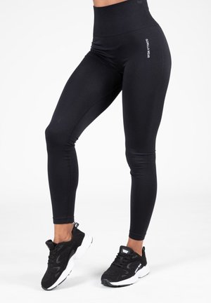 Zwarte high-waisted leggings van rekbare stof. Kenmerkt zich door een logo aan de zijkant en een gladde textuur. Gecombineerd met zwarte sportschoenen.