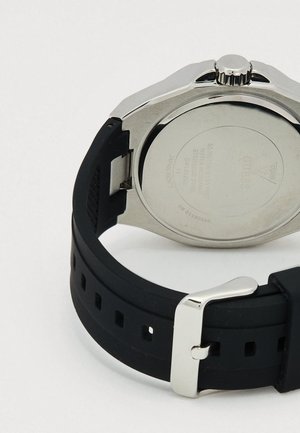 Zadaj strani uro Guess iz nerjavečega jekla s črnim silikonskim paščkom in srebrno zaponko, na kateri so prikazane podrobnosti o odpornosti na vodo in modelu.