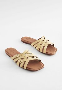 Brune flade sandaler med krydsede gule stropper, en firkantet tå og en lav hæl. Spænde detaljer på en strop. Glat tekstur.