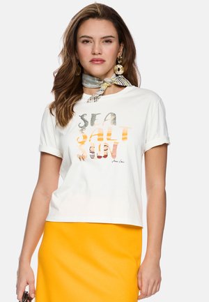Frau trägt weißes T-Shirt mit der Aufschrift "SEA SALT SUN", gelben Rock, gemustertes Halstuch und große runde Ohrringe vor weißem Hintergrund.