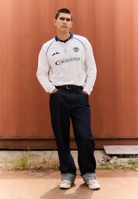 Hvit rugby-skjorte med lang erme og blå detaljer, kombinert med mørk denimjeans. Personen står med hendene i lommene foran en oransje bakgrunn.