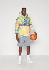 Hættebluse med et flerfarvet abstrakt print i grøn, gul og blå, parret med grå shorts og hvide sneakers, mens han holder en basketball.