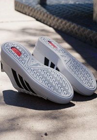 Zapatillas blancas con rayas negras colocadas con la suela hacia arriba sobre una superficie de concreto iluminada por el sol cerca de un cuerpo de agua.