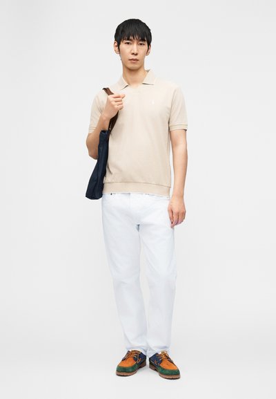 Άνδρας που φοράει μπεζ πόλο πουκάμισο, λευκό παντελόνι, πολύχρωμα παπούτσια και κρατά μια ναυτικό τσάντα ώμου σε λευκό φόντο.