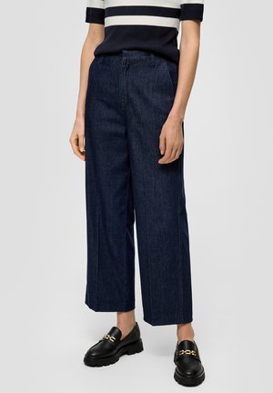 Persoon met donkerblauwe wijde cropped jeans, zwarte loafers met gouden kettingdetail, en een marineblauw-wit gestreept shirt met korte mouwen.