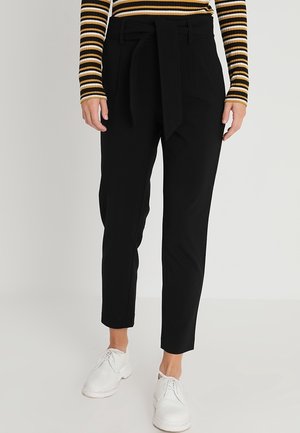 Personne portant un pantalon noir taille haute fuselé avec ceinture en tissu, un haut à manches longues rayé et des chaussures blanches à lacets, debout devant un fond uni.