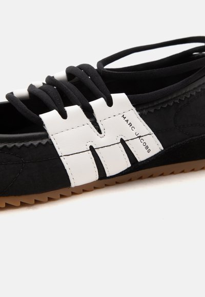 Gros plan d'une chaussure noire avec un porte-lacets en cuir blanc arborant la marque "MARC JACOBS" sur le côté et une semelle marron rainurée.