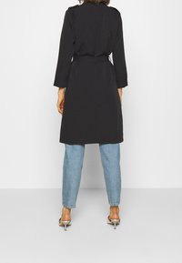 Trench-coat noir à ceinture avec manches longues, vue de dos. Associé à un jean droit bleu clair et des sandales à talons argentées.