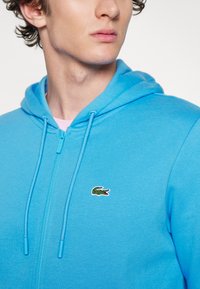 Sudadera azul con cremallera y cordones, hecha de algodón. Presenta un pequeño logo de cocodrilo verde en el pecho, textura suave.