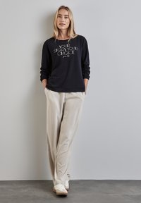 Schwarzer Sweatshirt mit weißem Text "DEINE GESCHICHTE. DEINE WAHL." Lässiger Schnitt. Beigefarbene Hose mit glatter Textur. Weiße Sneaker.