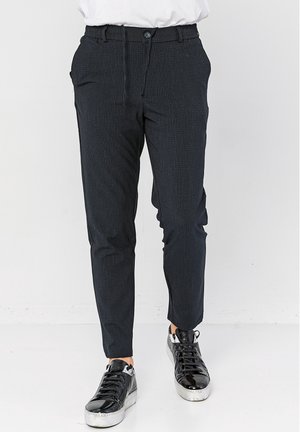 Homme portant un pantalon slim foncé et des baskets noires brillantes, debout sur un fond blanc uni.