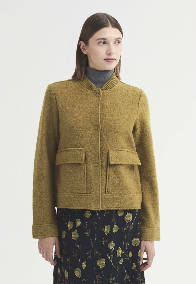 Femme portant une veste jaune moutarde avec des poches avant sur un col roulé gris et une jupe noire à fleurs, regardant sur le côté.