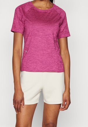 Kobieta ubrana w różowy melanżowy t-shirt z krótkim rękawem i okrągłym dekoltem, zestawiony z białymi szortami do kolan na jednolitym tle.