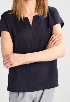 Person iført en mørkeblå kortærmet bluse og lysegrå bukser med armene krydset over brystet mod en ensfarvet baggrund.